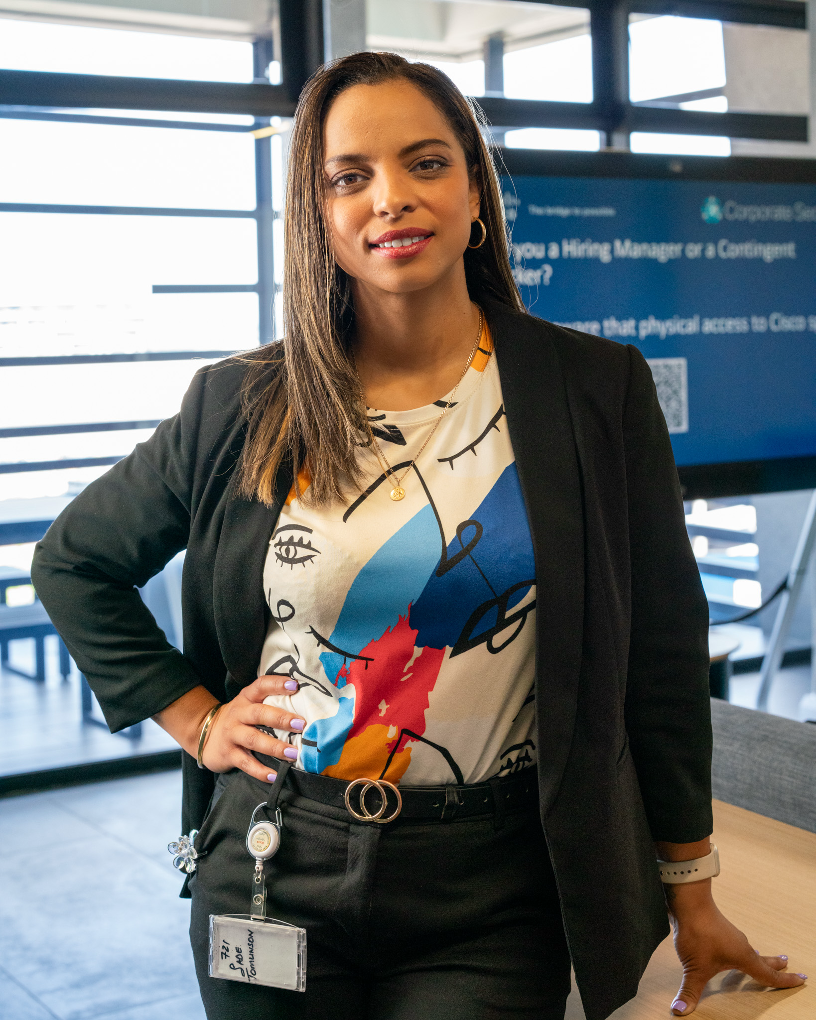 Woman in a black blazer and patterned top standing in an office space, wearing a badge and smiling at the camera.