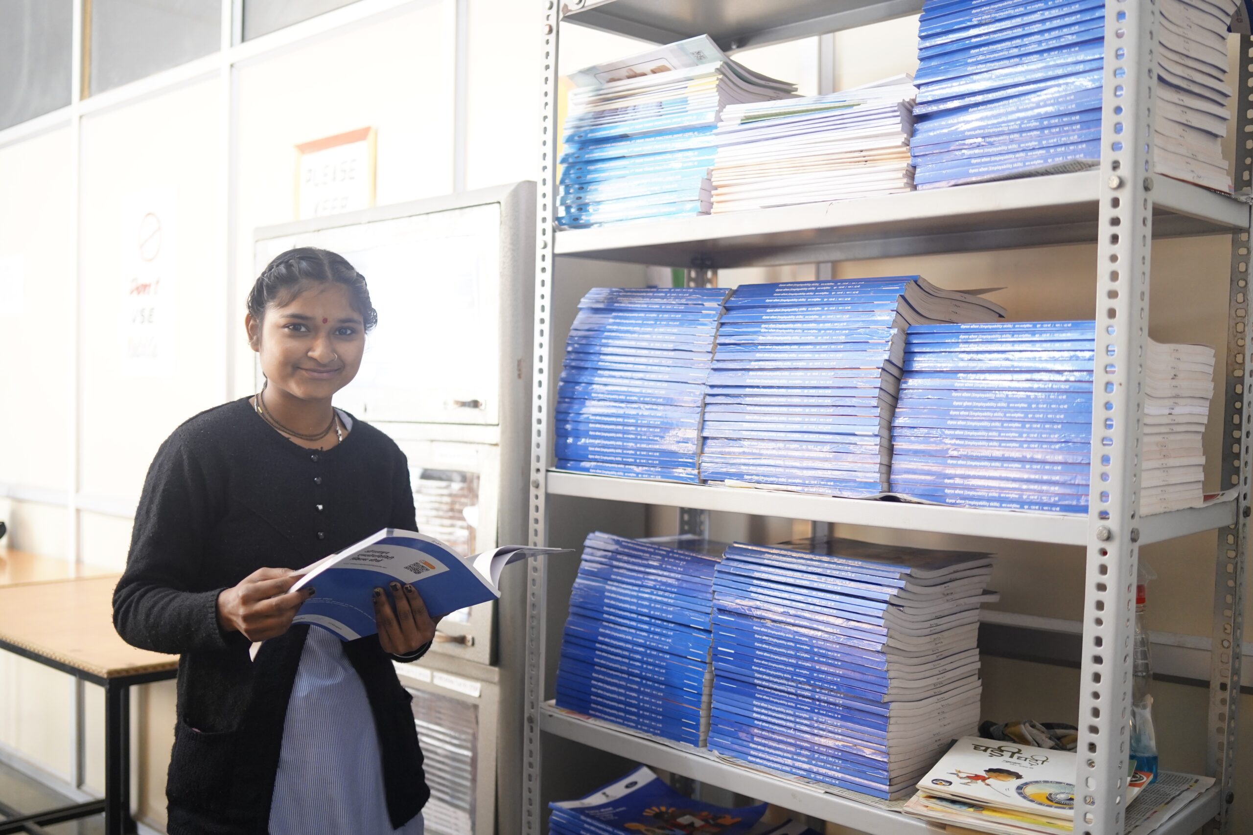 A young woman in India stands to the left of a big shelf with many stacks of blue textbooks. She is holding one of the open books in her hands.