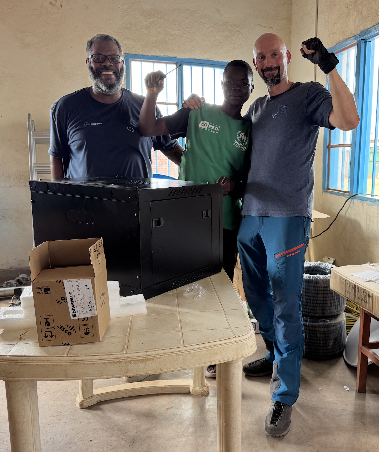 Three men stand in front of a piece of tech equipment in an office at the Musenyi refugee site in Burundi. All three men are smiling, and the men in the middle and to the right raise their fists in a sense of accomplishment.