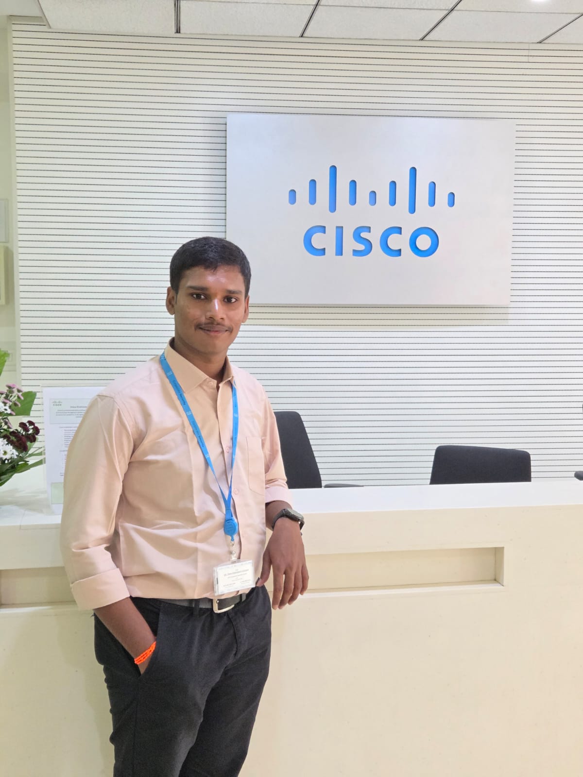 Young man with dark hair and light mustache stands, leaning one arm, across a white desk with a blue "Cisco" logo sign hanging on the wall in the background. 