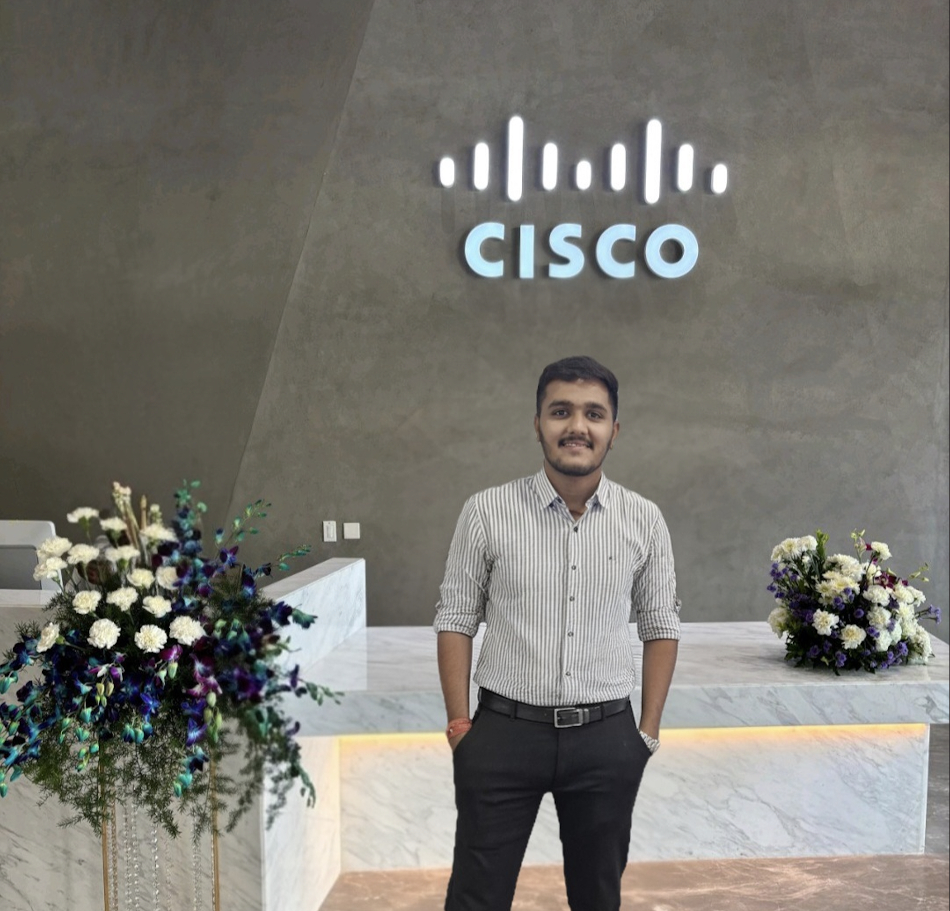 A man stands smiling in a modern office lobby beneath a glowing Cisco logo, with marble counters and floral arrangements on either side.
