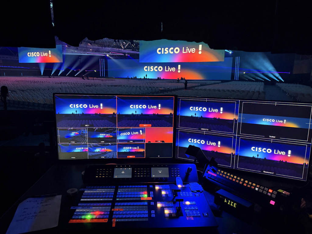 Production control room view of a large Cisco Live event, with multiple monitors displaying the stage and a mixing console in the foreground, overlooking a brightly lit stage and empty audience seating.