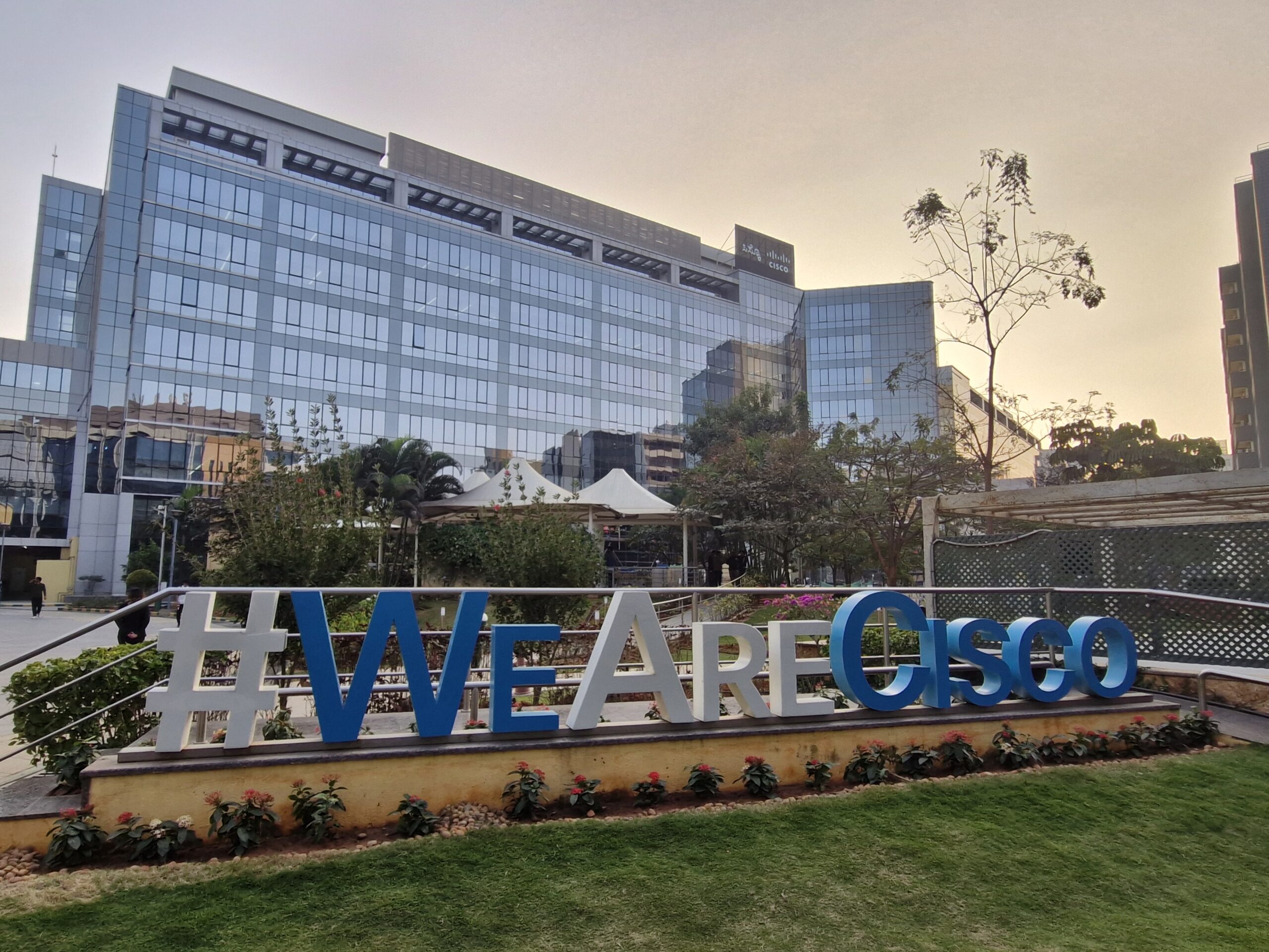 Exterior view of a modern glass office building at sunset with a large “#WeAreCisco” sign in front, surrounded by greenery.