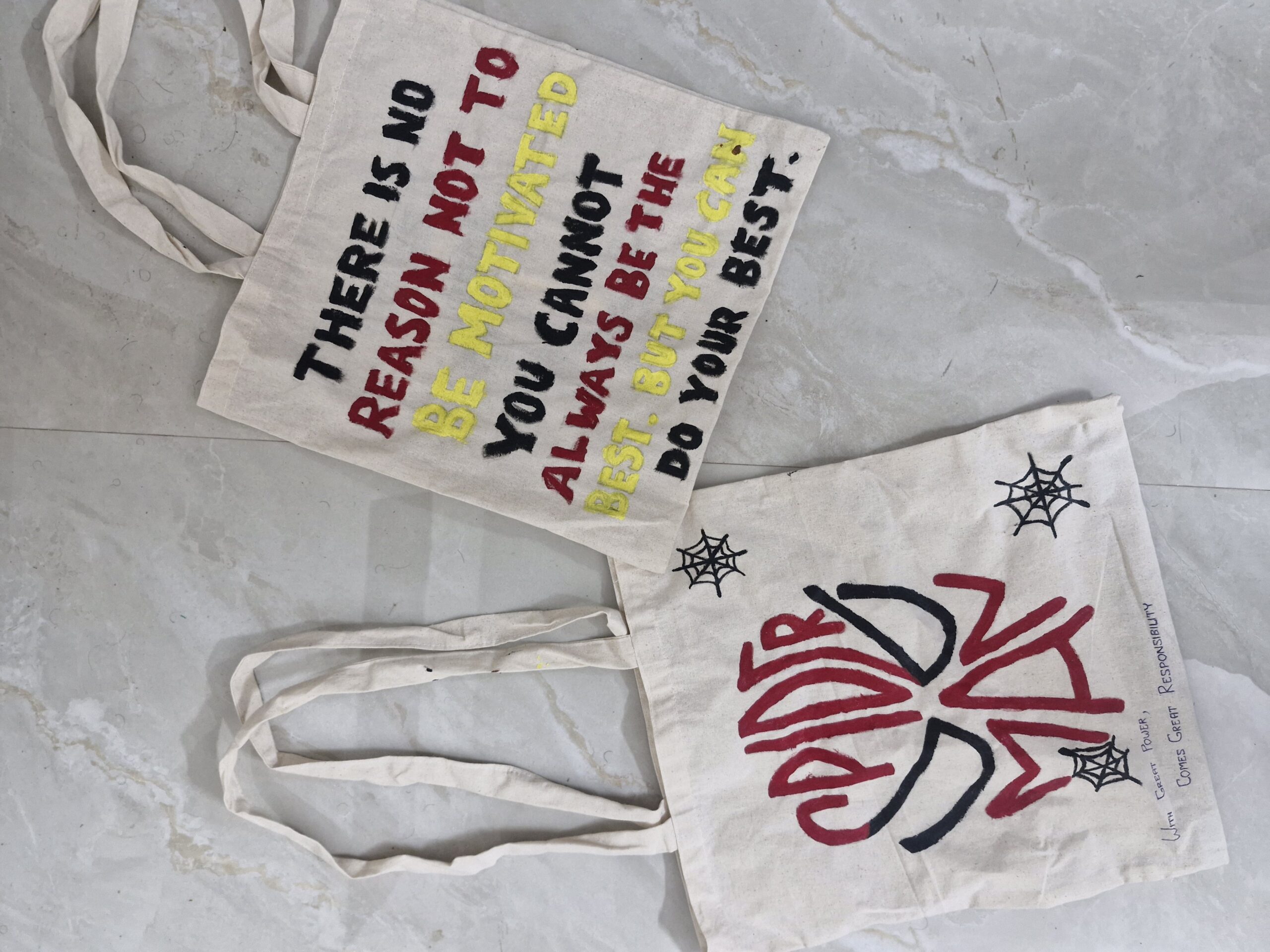 Two canvas tote bags on a tiled floor—one with a Spider-Man design and the other with a motivational quote in black, red, and yellow text.