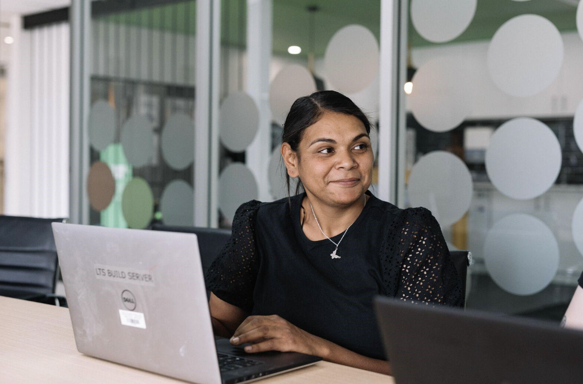 A young woman in a black blouse sits at a table in a conference room in front of a laptop.