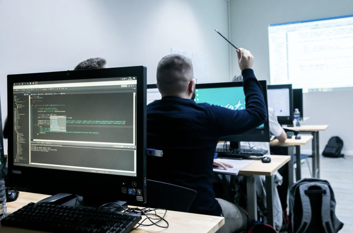 This picture is of a computer lab-style classroom. To the left is a large computer monitor. In the middle, a man (from the back) seated in from of a computer raises his hand.