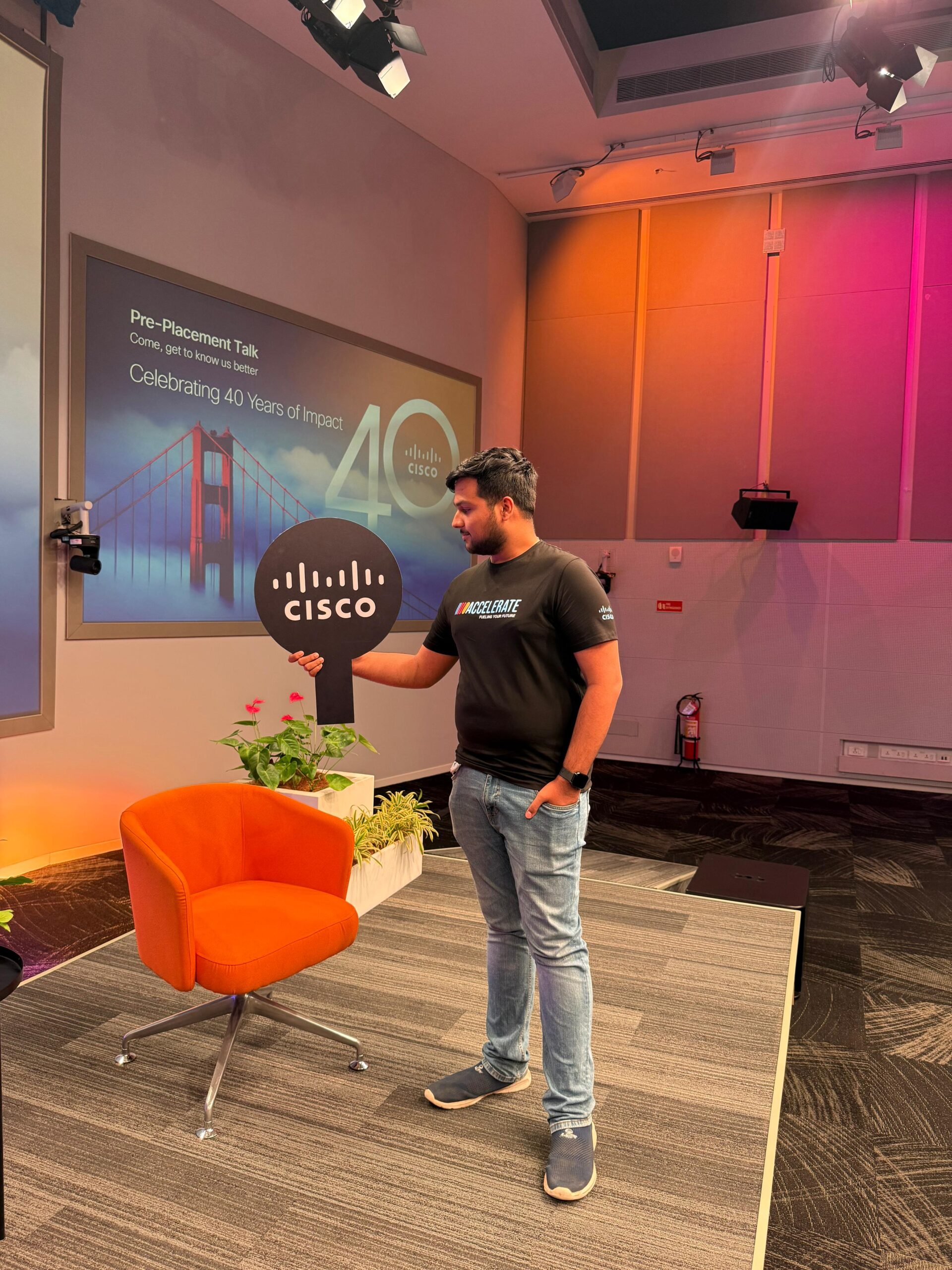 Man in black "Accelerate" t-shirt stands near an orange chair, holding a black sign with Cisco logo. A Cisco 40th anniversary screen featuring the Golden Gate Bridge is in the background.