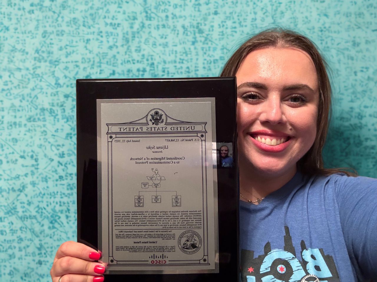Woman smiling while holding a framed Cisco patent plaque against a teal patterned background.