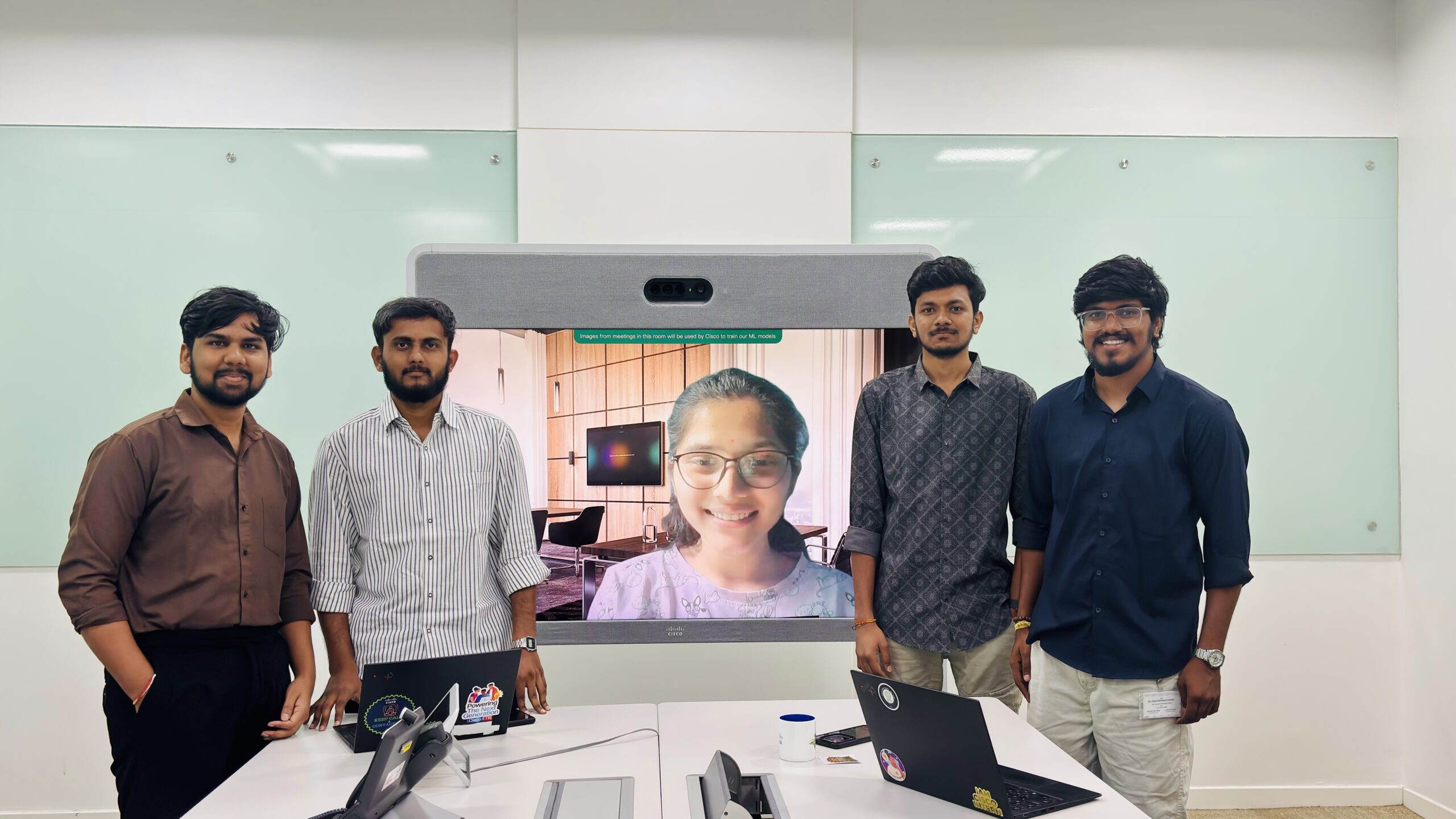 Four Cisco volunteers stand at a table with laptops during the Tech for Social Good Hackathon, presenting their work on a monitor to a remote team member displayed on screen. The team is in a modern office space with mint green glass walls.