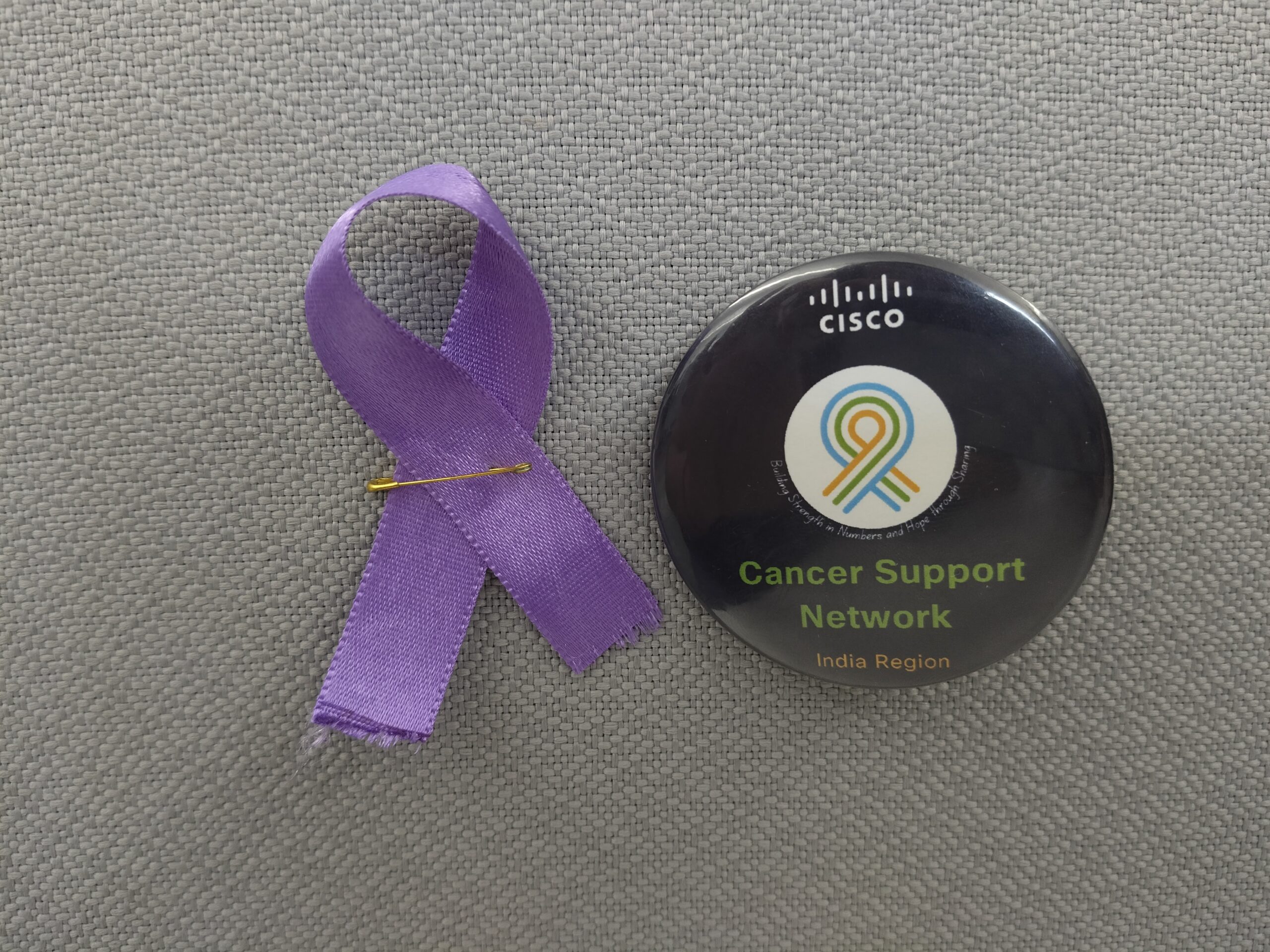 A purple awareness ribbon pinned beside a Cisco Cancer Support Network button labeled “India Region,” placed on a gray fabric background.