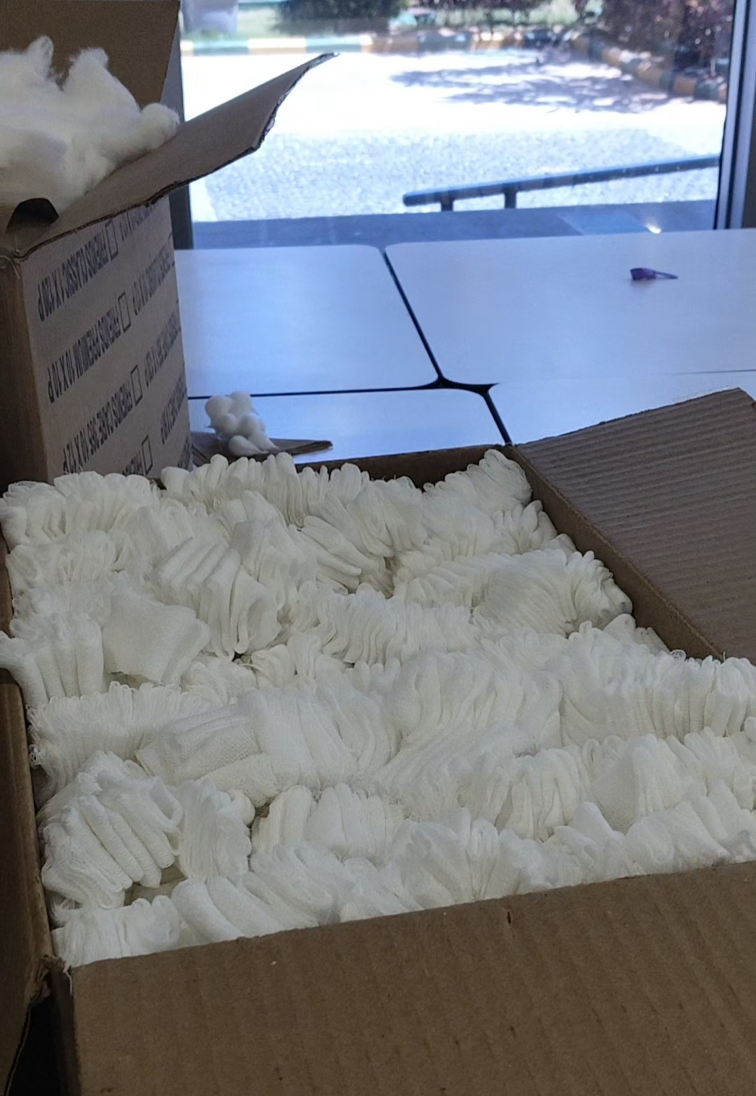 Open cardboard boxes filled with neatly stacked white fabric items or gloves on a table near a window, with packing material visible in the background.