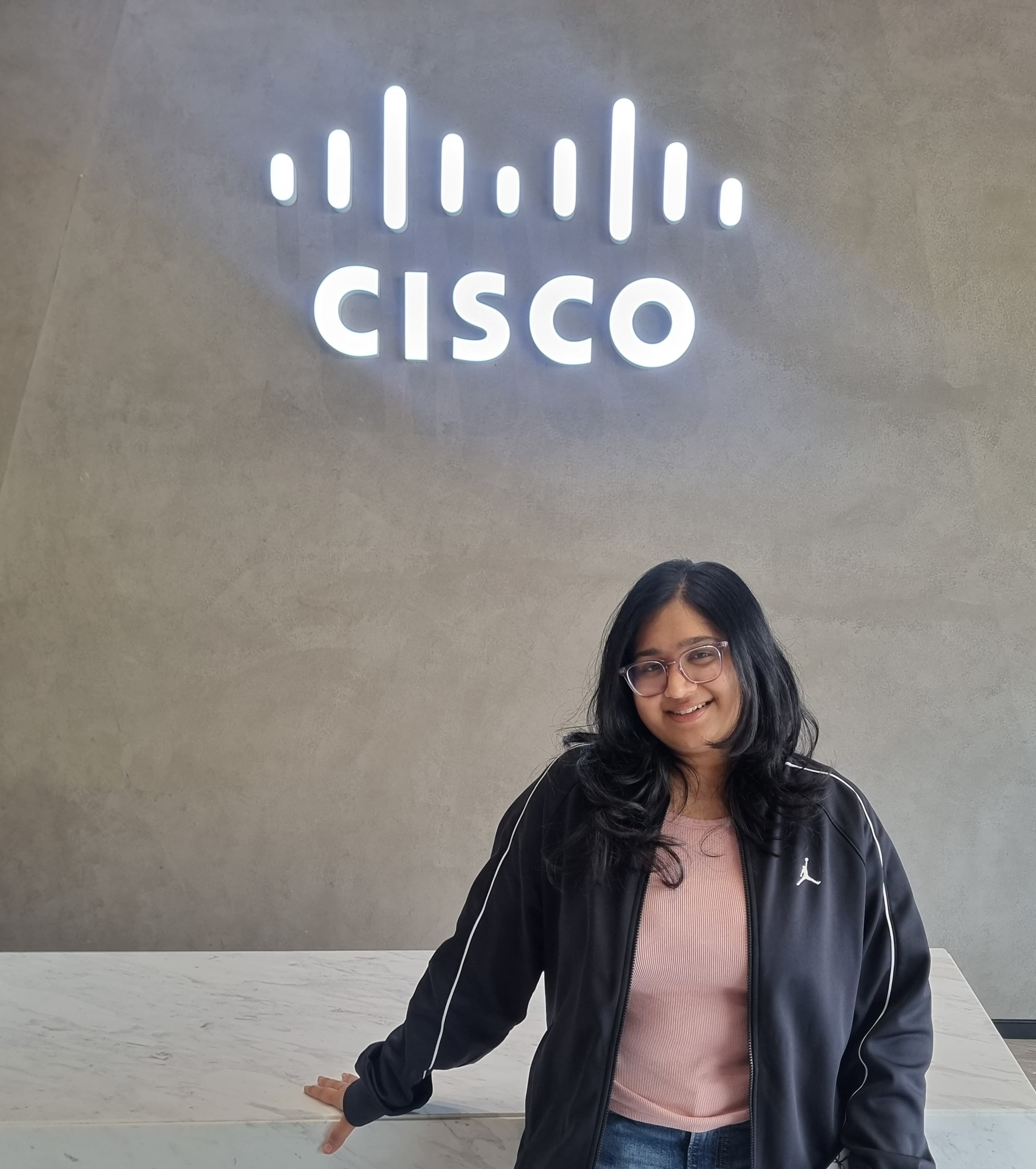 Young woman with dark hair, wearing a pink shirt and black jacket, stands in front of a wall with an illuminated "CISCO" logo. 