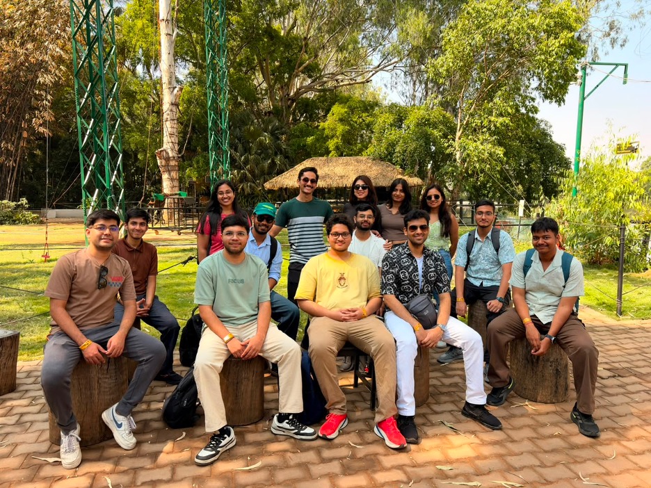 A group of interns pose outdoors in a landscaped park setting. Some are seated on tree-stump benches while others stand behind them. The background includes green trees, walking paths, and adventure-park structures. The group is smiling and dressed casually, enjoying a team outing on a sunny day.