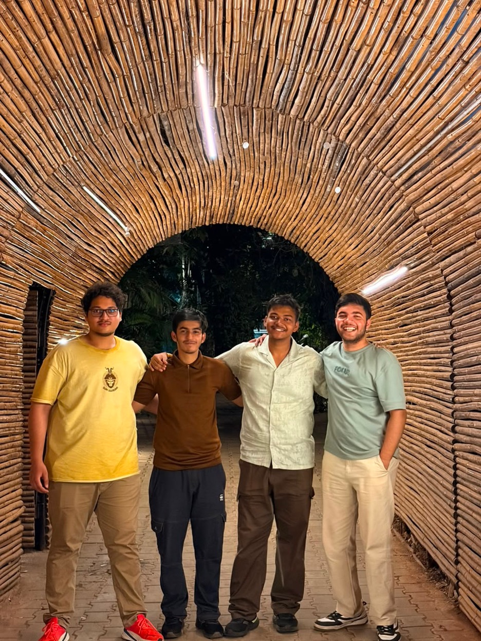 Four interns stand together beneath a curved tunnel made of bamboo or wooden poles. The tunnel creates a dramatic archway framing the group. The interns have their arms around each other and are smiling at the camera. Warm lighting and natural textures give the photo a friendly and relaxed feel.