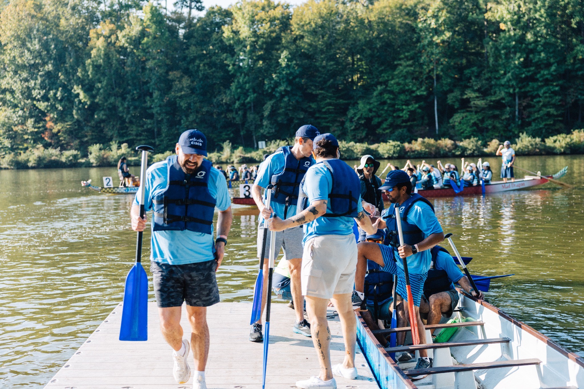A group of employees wearing matching light-blue shirts, life jackets, and caps prepare for a dragon boat race on a lake. Several team members board a long canoe-style boat from a dock while holding paddles. Additional dragon boat teams are visible on the water in the background, surrounded by trees on a sunny day. The scene captures teamwork, collaboration, and outdoor employee engagement.
