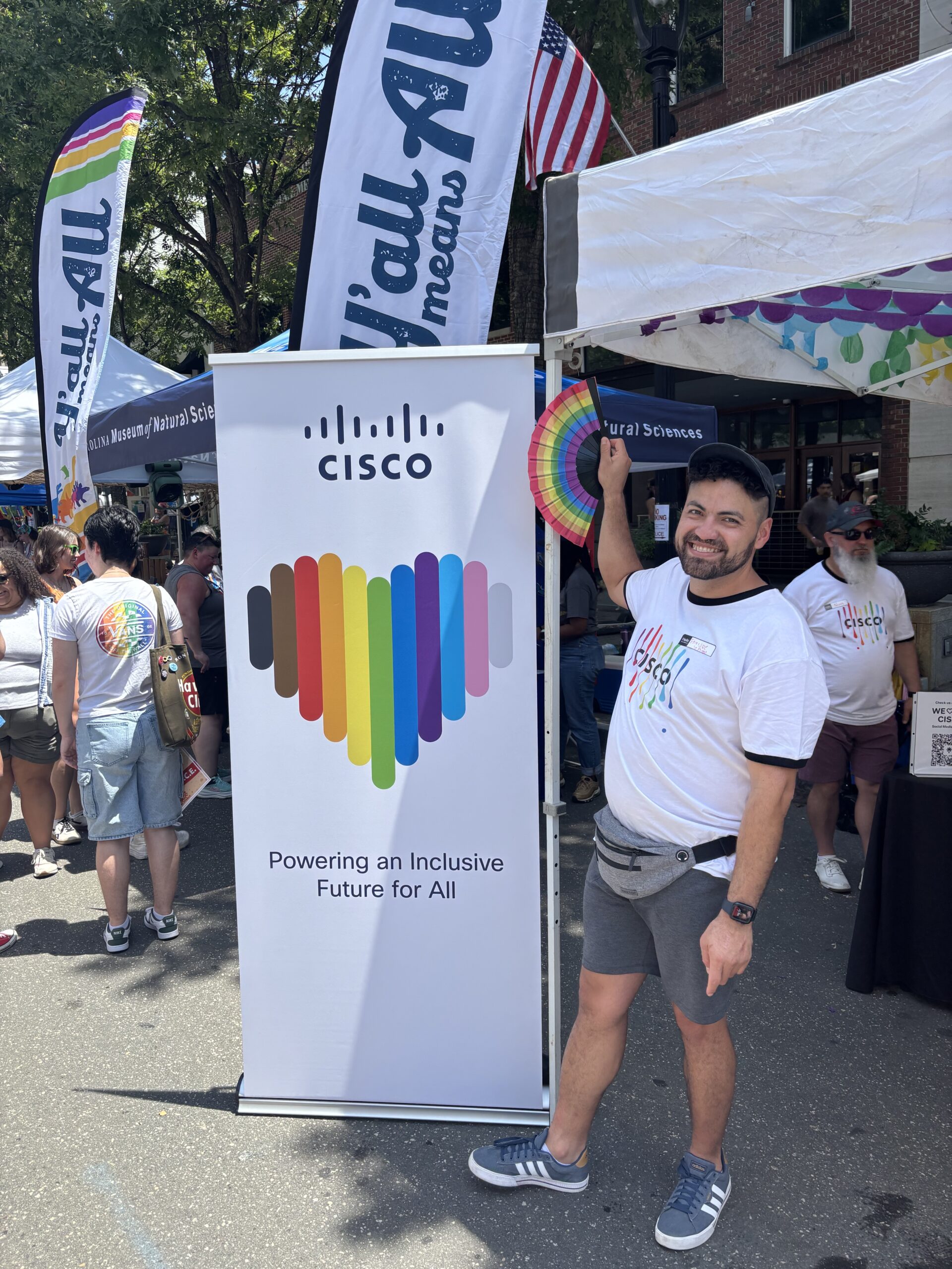 A smiling Cisco employee stands beside a Cisco-branded banner featuring a rainbow-colored heart and the message “Powering an Inclusive Future for All.” The photo is taken at an outdoor community festival with tents, flags, and attendees visible in the background.