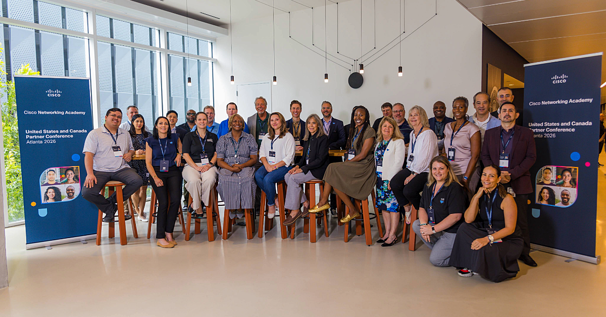 30-35 people stand together in a group photo posing between two signs at the 2026 Cisco Networking Academy Partner Conference for United States and Canada partnerships.