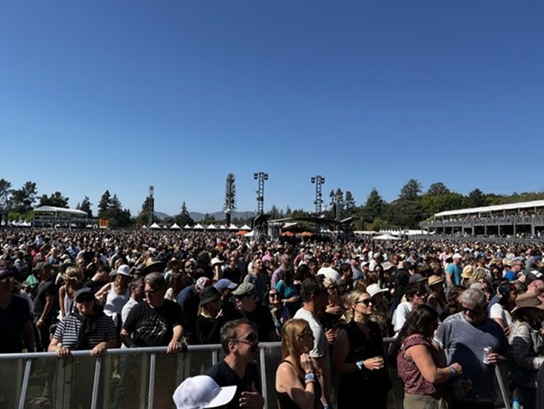 A large crowd gathered on a sunny day at an outdoor music festival, with a stage visible in the background.