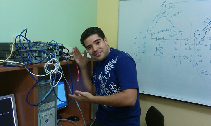 Standing beside networking equipment in a classroom lab, pointing to routers and cables.