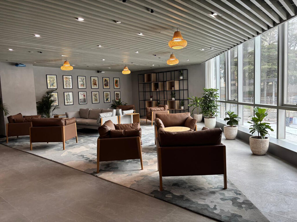 A comfortable office lounge area featuring brown leather armchairs, a grey sofa, and a large patterned area rug, accented by woven pendant lights, indoor plants, and a gallery wall of framed art.