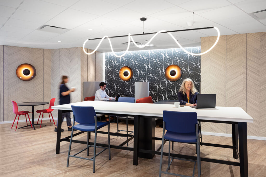 A modern, vibrant office cafe area with a large white table, blue chairs, a geometric patterned wall, and a distinctive, flowing circular light fixture overhead.
