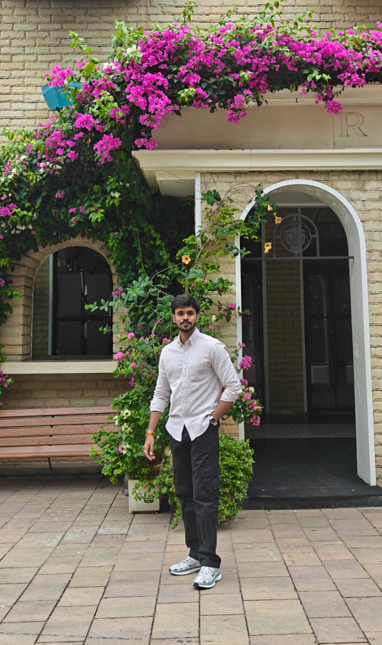 A professional headshot of a man with dark hair and beard, wearing a light-colored button up shirt, standing outdoors in front of a wall covered in lush green vines and bright pink bougainvillea flowers.