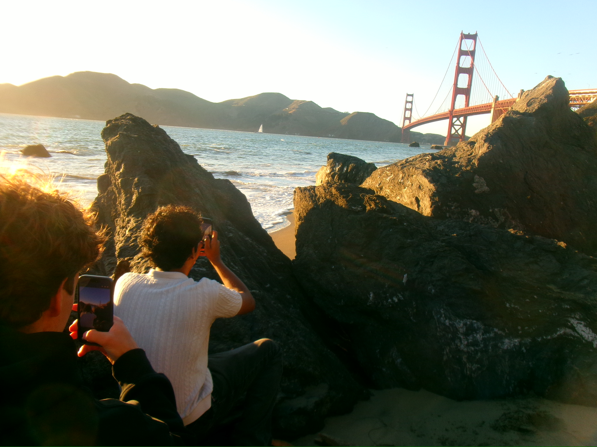 Two people photograph the Golden Gate Bridge from large coastal rocks along a sandy beach during golden hour.
