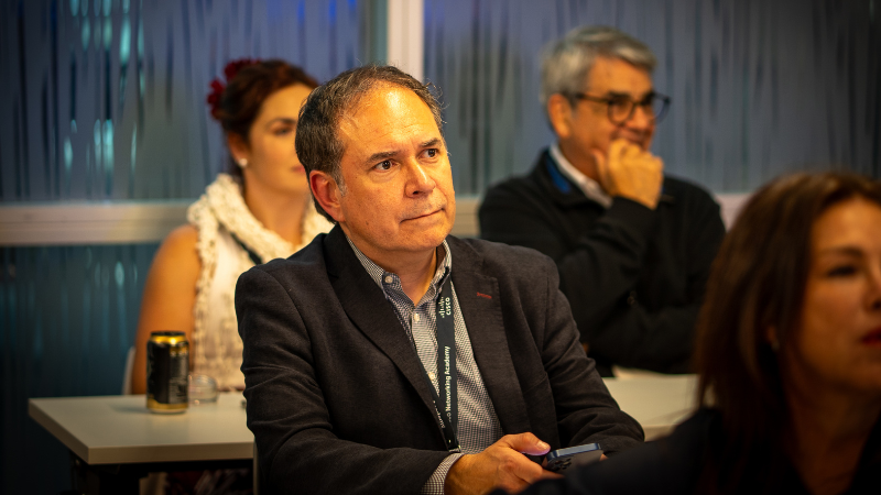 A man sitting in the audience, holding a smartphone and looking focused toward the front of the room during a presentation.