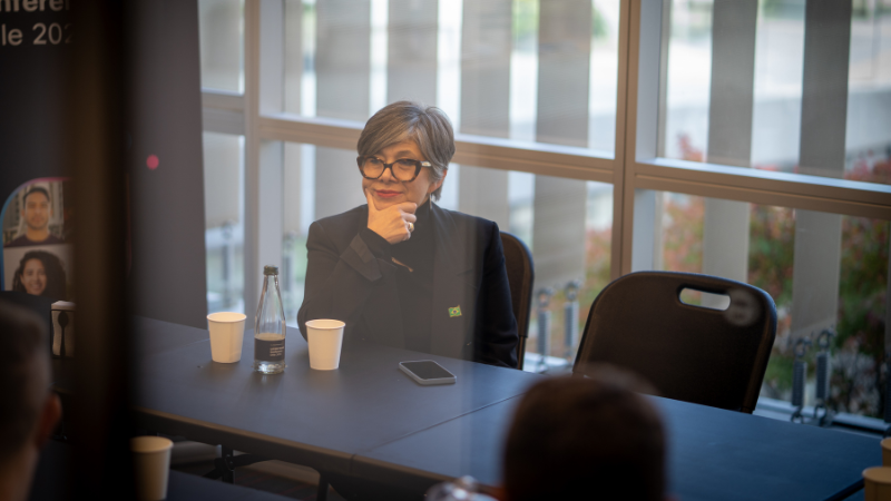 A woman with short grey hair and glasses sitting at a table, listening attentively during a session at the Cisco Networking Academy LATAM Partner Conference.