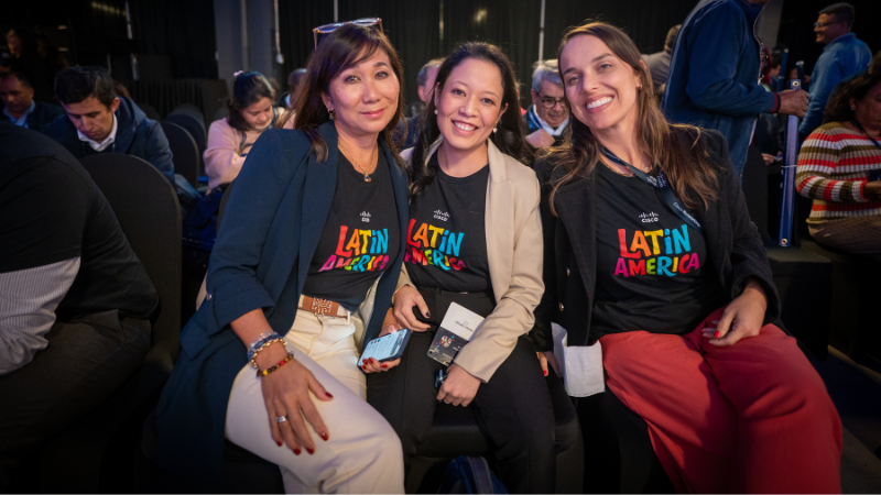 Three women smiling at the camera. Two are wearing black t-shirts featuring the words "LATIN AMERICA" in colorful typography.