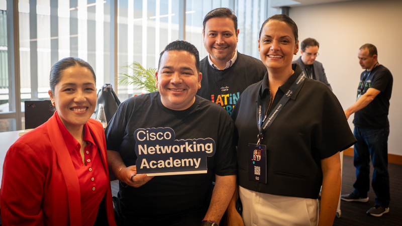Four attendees smiling for a photo, with one person in the center holding a blue "Cisco Networking Academy" sign.