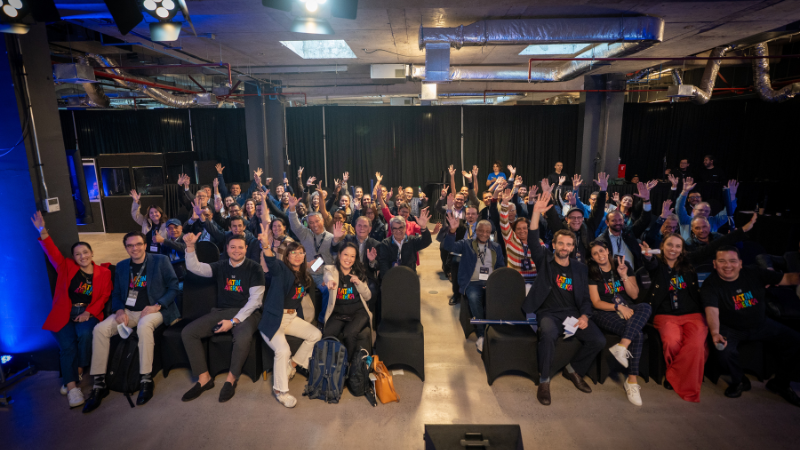 A large audience of attendees sitting in rows of chairs at the conference, raising their hands and smiling enthusiastically for the camera.