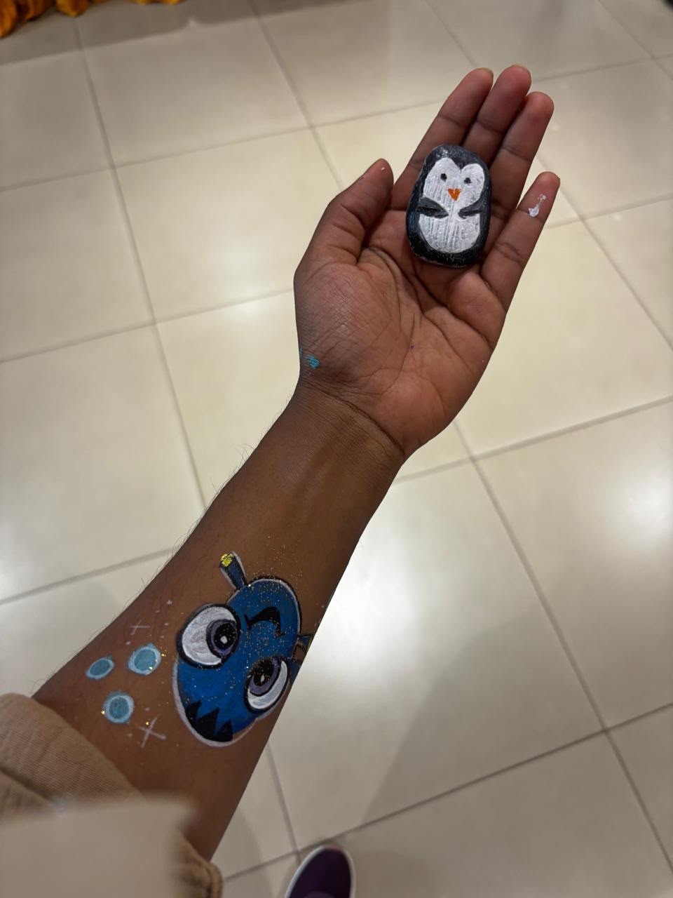 A person holds a small hand-painted penguin rock in their palm, with a colorful blue character painted on their forearm.