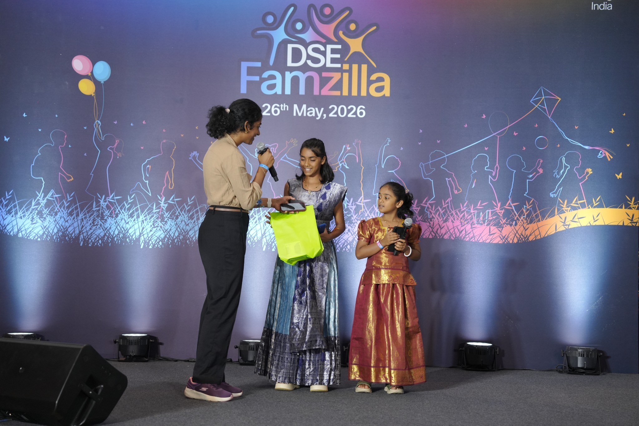 A woman on stage hands a green gift bag to a girl in a blue traditional dress, while another girl in a red and gold traditional dress stands nearby holding a microphone. The backdrop features a "DSE Famzilla" event banner dated May 26, 2026.