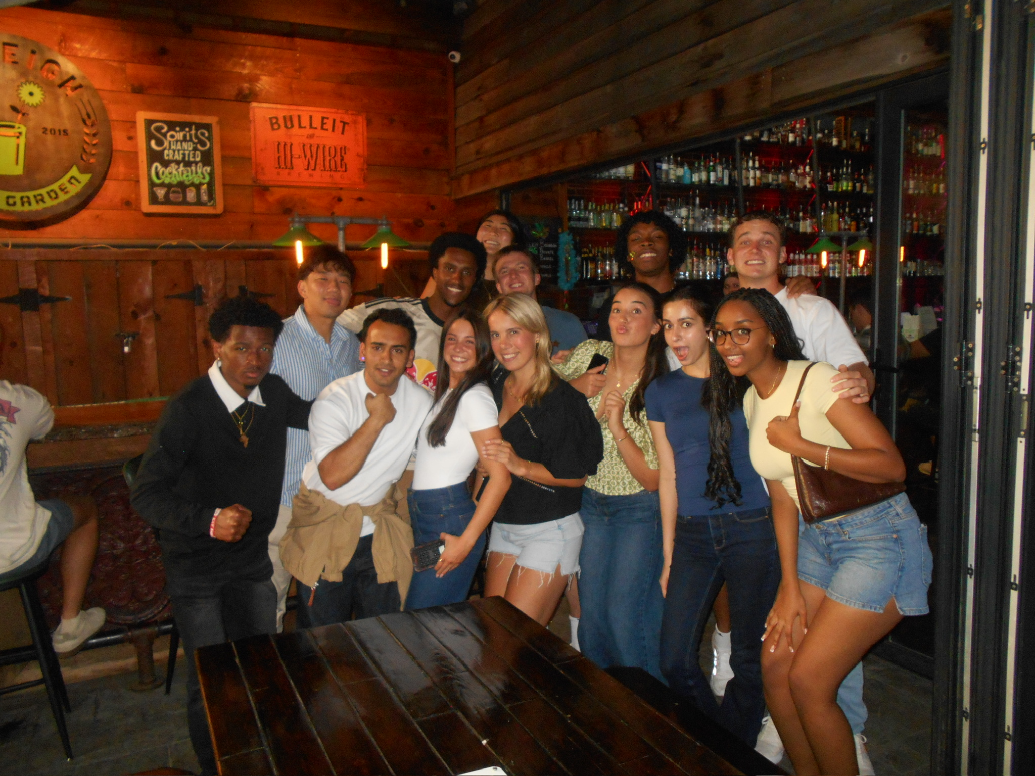 A group of young adults gathers closely together for a group photo inside a rustic restaurant with wooden walls and warm lighting.