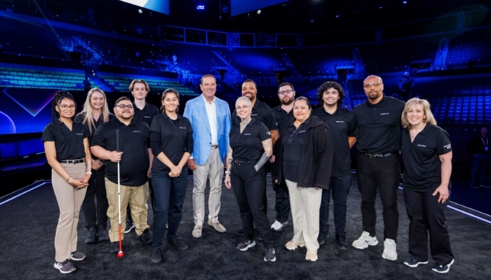 Thirteen Cisco Networking Academy Dream Team members in black Cisco polos stand with Cisco CEO Chuck Robbins on a blue-lit arena stage at Cisco Live US 2026.