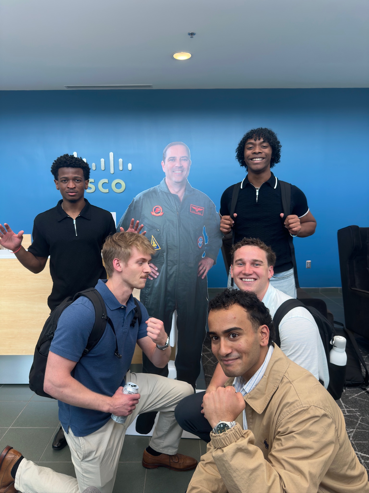 Five interns pose playfully around a life-size cardboard cutout in a Cisco office lobby with a blue branded backdrop.