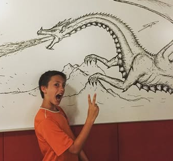 Boy in orange shirt holds up peace sign in front of dragon drawing on white board. 