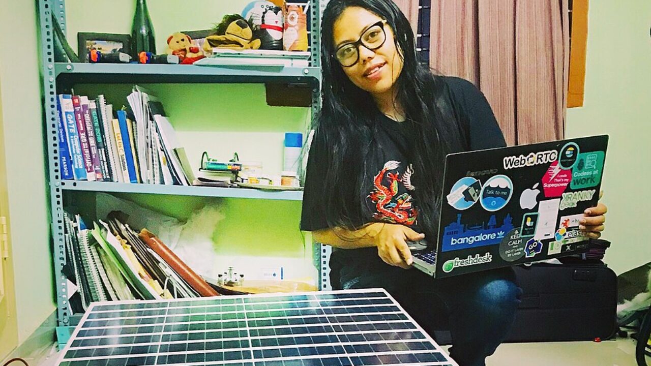 Woman sitting beside several small wheeled robot prototypes, holding a laptop covered with technology-themed stickers.