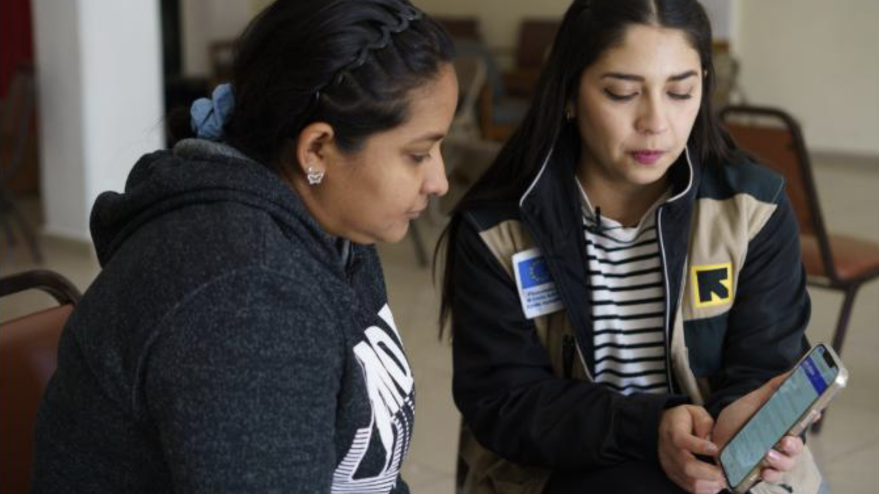 A woman in a sweatshirt sits next to a female caseworker wearing an IRC staff vest; the caseworker is holding a phone with the Signpost app/tool on screen.
