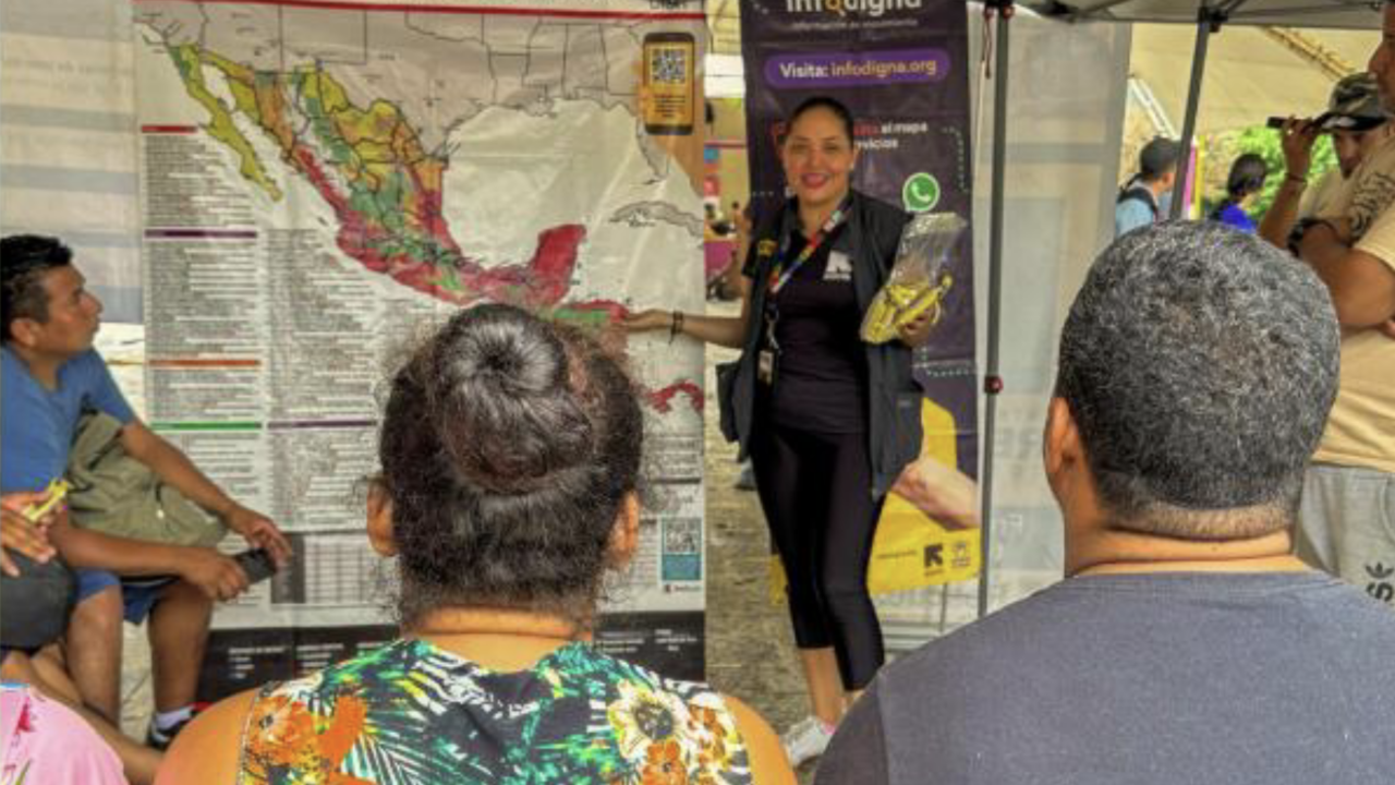 A female IRC staff member stands next to a banner/map in front of a small crowd, presenting information about the IRC's Signpost tool.