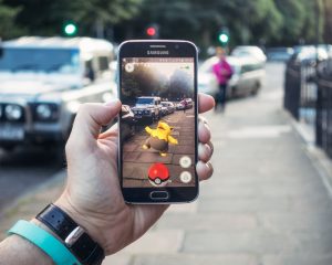 Edinburgh, UK - July 18, 2016: Closeup of a man holding a Samsung S6 smartphone, playing Pokemon Go with the game's augmented reality superimposing a character onto the pavement surface, as a person approaches in the distance.